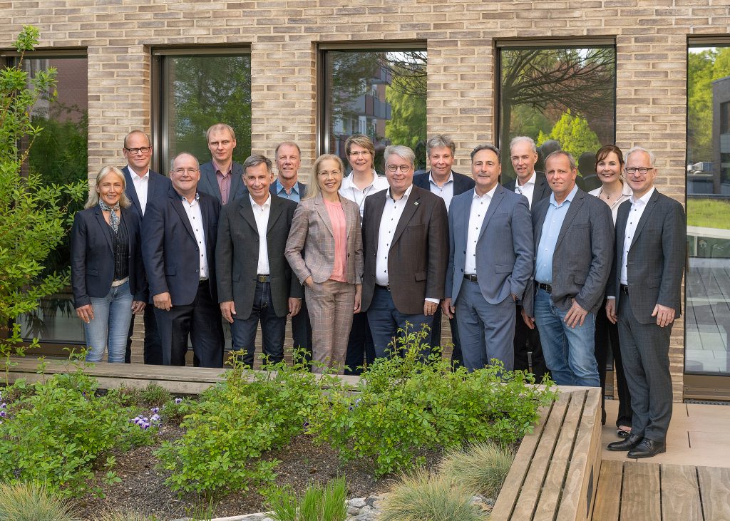 15 Personen in Businesskleidung stehen vor einem verklinkerten Gebäude mit vielen Fenstern. Vor Ihnen befindet sich ein kleiner Garten. Die Personen lächeln freundlich in die Kamera.