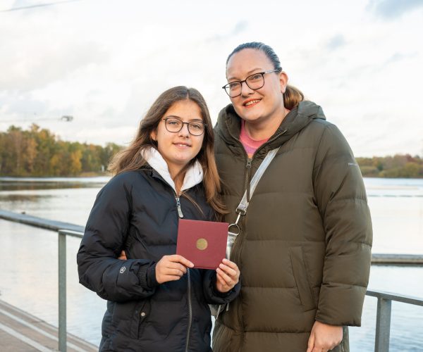 Ein Mädchen und eine erwachsene Frau stehen nebeneinander an einer Promenade und lächeln in die Kamera. Das Mädchen hält ein Zertifikat in der Hand.