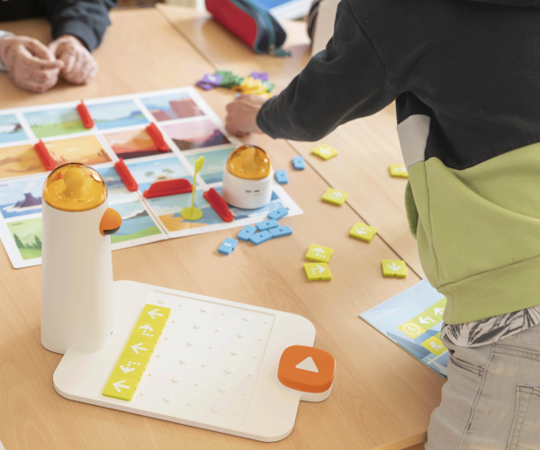 AUf einem Schultisch liegt ein bunter Plan und verschiedene bunte Plastikchips, die darauf angeordnet sind. Davor steht ein weiß-orangener Miniroboter.