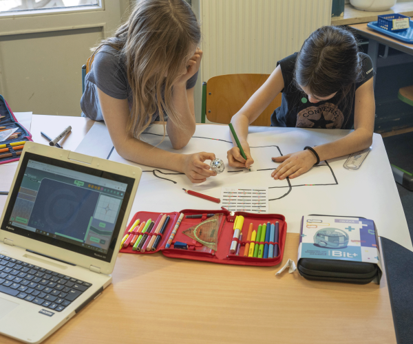 Eine Schülerin und ein Schüler sitzen an einem Schultisch über einer Zeichnung, auf der eine Fahrbahn verläuft. Auf dem Tisch stehen außerdem ein geöffnetes Federmäppchen und ein Laptop.