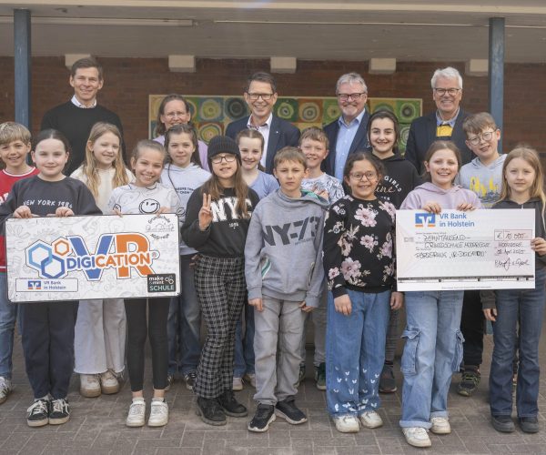 Kinder und Erwachsene stehen vor einem Schulgebäude. DIe Kinder in der ersten Reihe des Fotos halten einen großen Scheck sowie eine Grafik mit dem VR-Digication-Logo.
