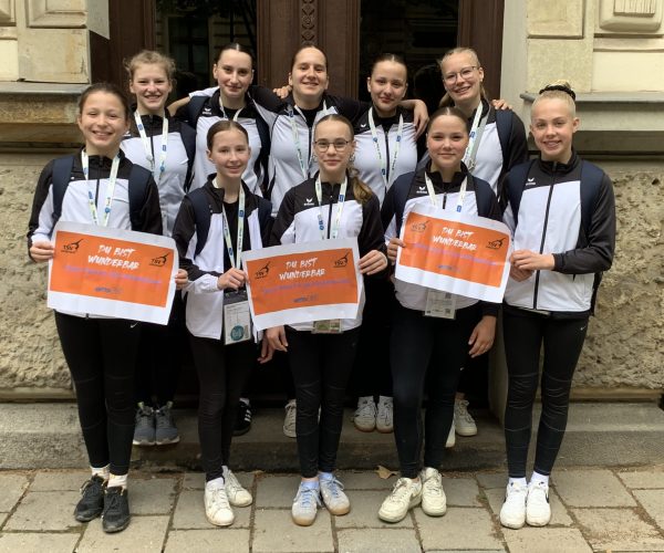 Eine Gruppe von Mädchen in schwarz-weißen Trainingsanzügen steht aufgereiht vor einem Gebäude. Die Mädchen in der ersten Reihe halten zwei Schilder, auf denen in blau-orangener Schrift steht "du bist wunderbar"